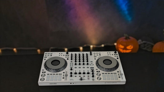 White Pioneer DJ DDJ-FLX10 controller on a dark booth table, with colored stage beams, haze, string lights and a carved pumpkin behind it at a Halloween party.